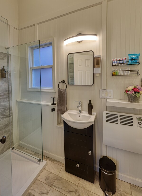 Une salle de bain avec une douche à parois de verre, un lavabo blanc avec un miroir au-dessus et une poubelle noire.