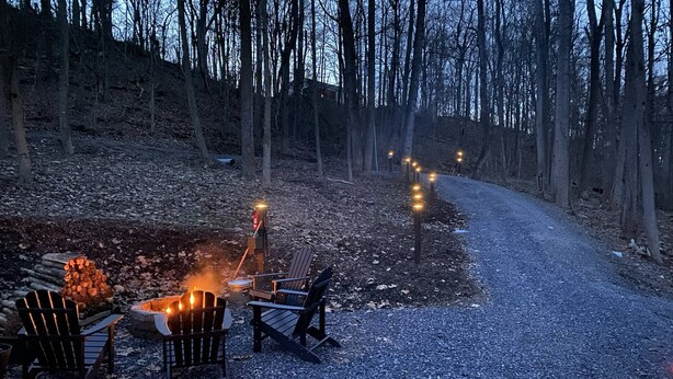 A gravel path leading through a wooded area with a fire pit and chairs set up for outdoor seating.