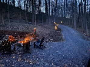 Un chemin de gravier traverse une zone boisée où se trouvent un foyer et des chaises disposées pour créer un espace de détente en plein air.
