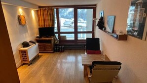 Un salon chaleureux avec un plancher en bois, un téléviseur, une cheminée et une grande fenêtre donnant sur un paysage enneigé.