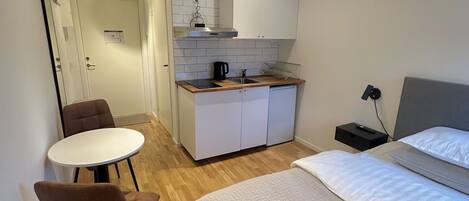A compact hotel room with a bed, a small kitchenette, a dining area with a table and chairs, and a bathroom visible in the background.