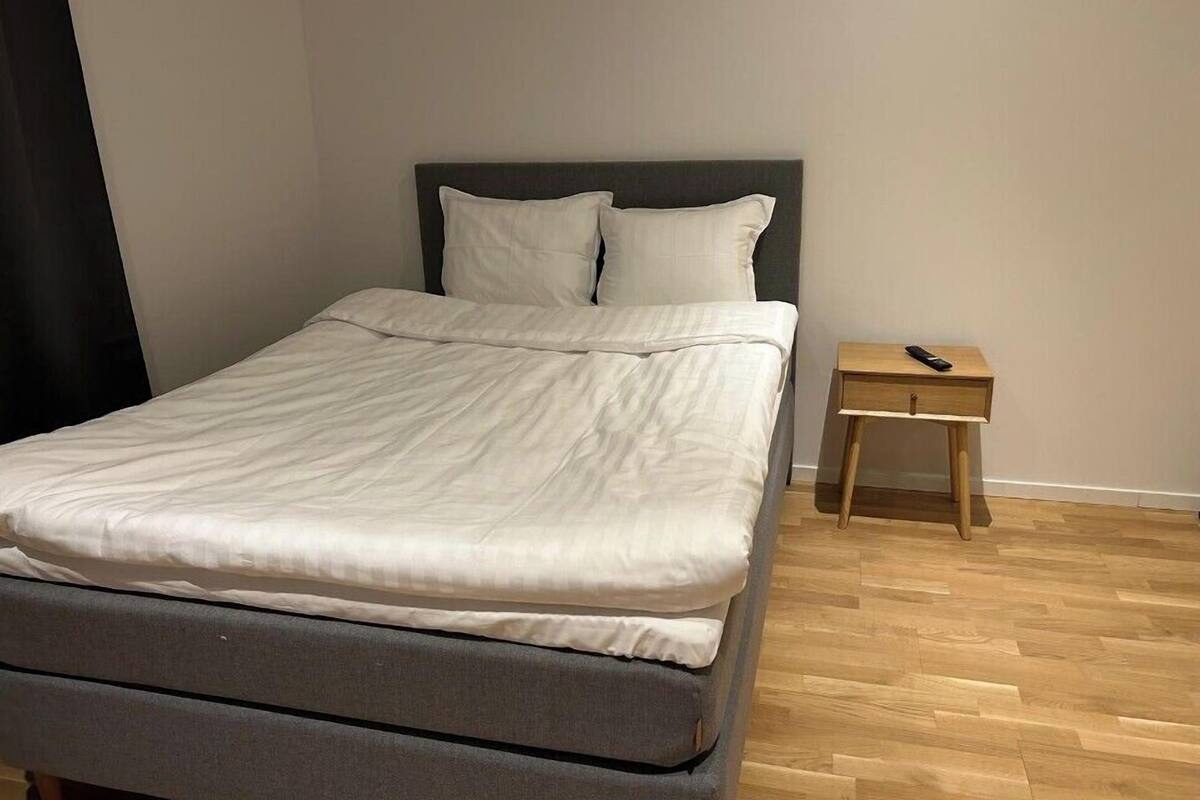 Een netjes opgemaakt bed met een grijze matras en wit beddengoed in een kamer met houten vloeren en een klein nachtkastje ernaast.