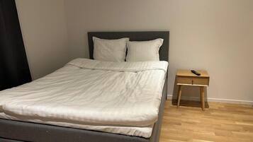 Una cama bien hecha con un colchón gris y ropa de cama blanca en una habitación con suelo de madera y una mesita de noche de madera al lado.