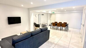 A modern hotel lobby with a grey sectional sofa, a flat-screen TV, a dining area with brown chairs, and a tiled floor. - Edsviken Hotell AB (Sollentuna)