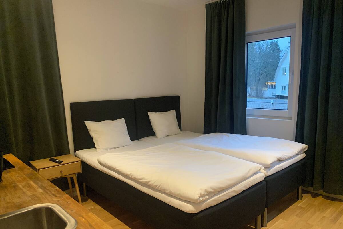 Een tweepersoonsbed met witte beddengoed, een houten nachtkastje met een telefoon, een klein wastafeltje en een raam met gordijnen.