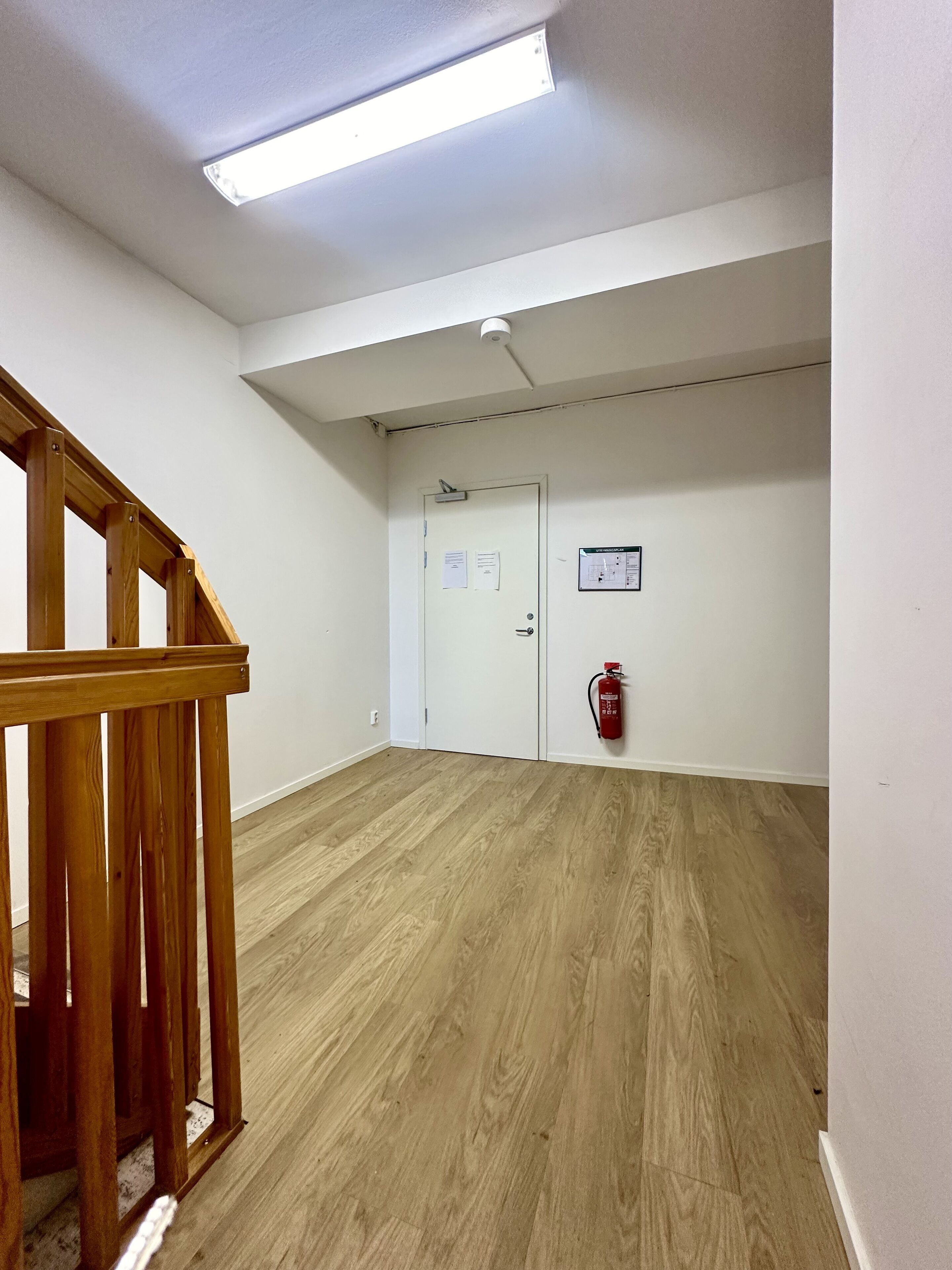 A spacious room with wooden flooring, a white door, and a fire extinguisher mounted on the wall.