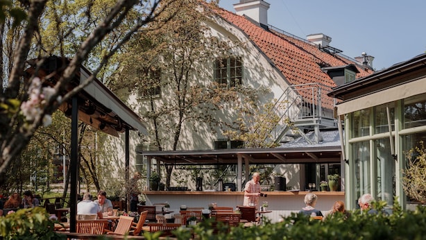 An outdoor café with wooden tables and chairs, a covered seating area, and a building with a tiled roof in the background. - Edsviken Hotell AB (Sollentuna)
