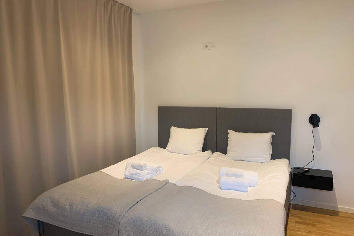 Een opgemaakt bed met een grijze deken, witte kussens en opgerolde handdoeken ernaast.