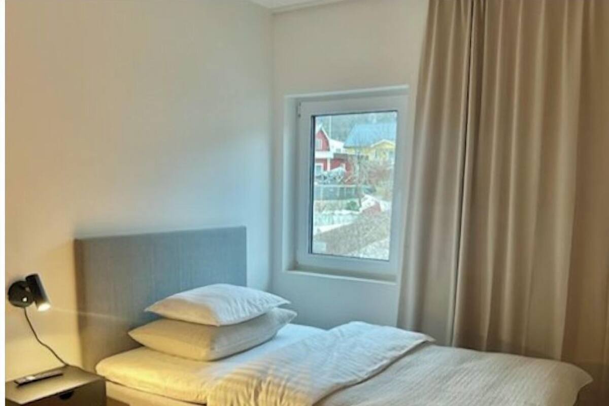 Een slaapkamer met een bed, een nachtkastje met een lamp, een raam met uitzicht op huizen en een gordijn.