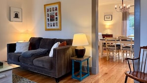 A living room with a dark brown sofa, a small table with a lamp, a framed collage on the wall, and a dining area with wooden furniture. - Cozy Country Getaway in the Heart of Upperville  (Upperville)