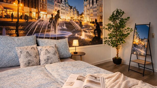 Un dormitorio con cama, almohadas, una mesita de noche, una lámpara, una planta en maceta y un mural con vista a la ciudad.