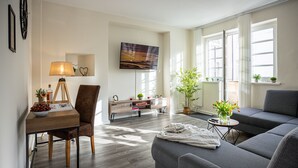 Una sala de estar moderna con un sofá, una mesa de comedor con sillas, un televisor montado en la pared y una ventana con plantas en el alféizar.
