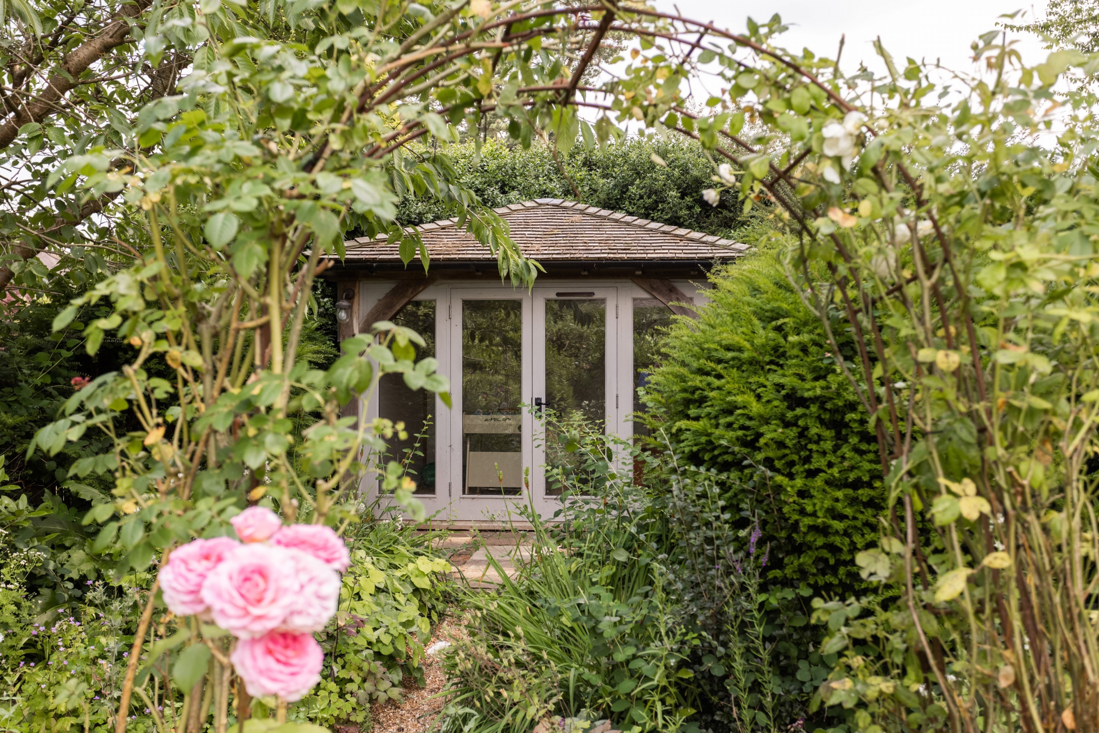 Une petite maison de jardin rustique, dotée d’un toit en pente, entourée d’une végétation luxuriante et de plantes en fleurs.