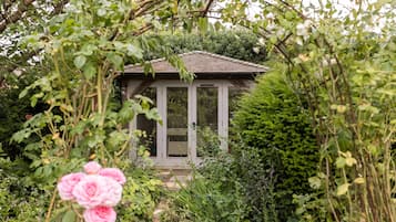 Une petite maison de jardin rustique, dotée d’un toit en pente, entourée d’une végétation luxuriante et de plantes en fleurs.