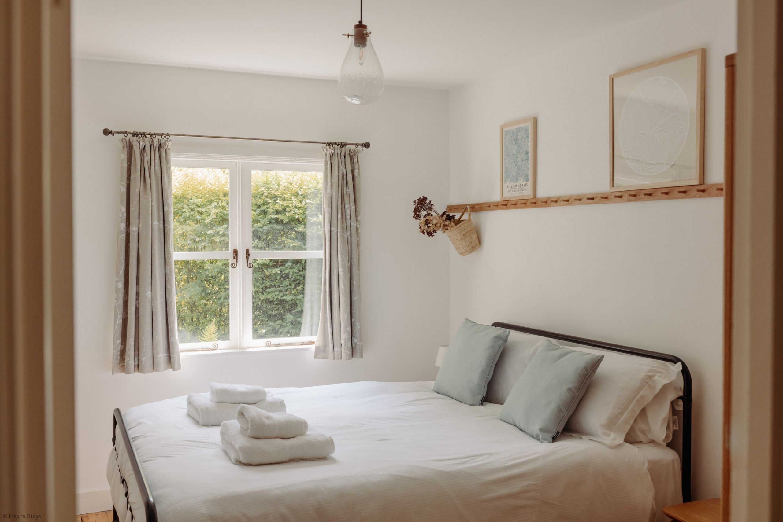 A bedroom with a bed, curtains, a hanging light fixture, a window with a view of greenery, and a shelf with a framed picture and a basket.