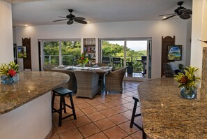 A kitchen with a granite countertop, bar stools, a dining table with chairs, a ceiling fan, and a view through large sliding glass doors to a balcony.