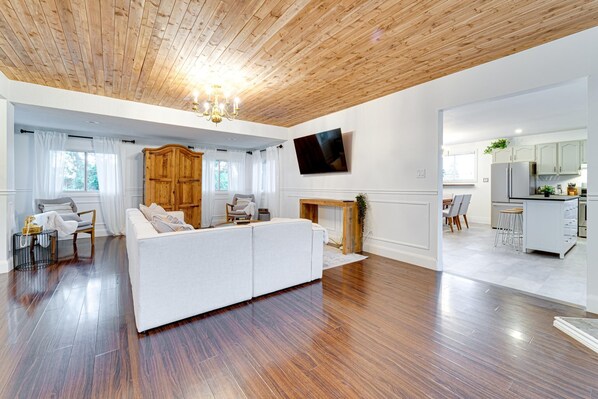 A modern living room with a wooden ceiling, a white sofa, and a TV mounted on the wall. - BOUTIQUE WINE COUNTRY HOME WITH POOL AND HOT TUB (Niagara-on-the-Lake)