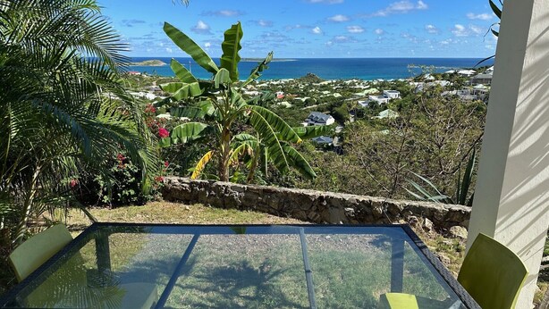 A table with chairs, a stone wall, and a view of the ocean and houses. - Villa Oasis Garden Apartment (Collectivity of Saint Martin)