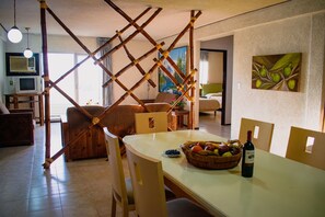 Uma área de refeições com uma mesa posta para dois, uma cesta de frutas e uma garrafa de vinho.