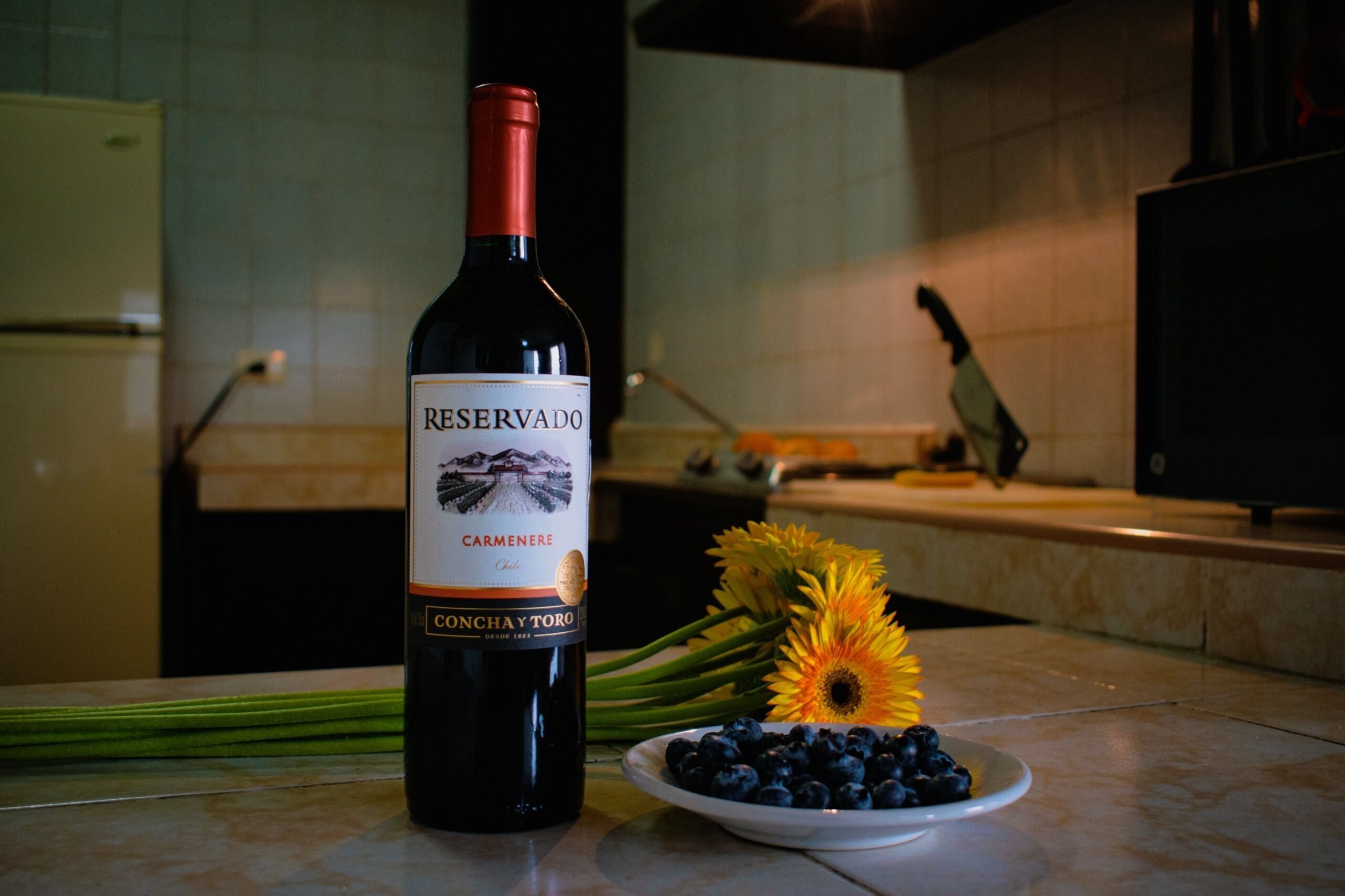 Una botella de vino Reserva Carmenere, un plato de arándanos y girasoles sobre una encimera de cocina.