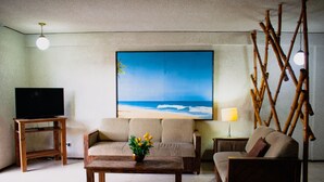 A living room with a large beach-themed painting, a television on a wooden stand, a sofa, a coffee table with flowers, and a lamp. - Suites del Sol (Mérida)