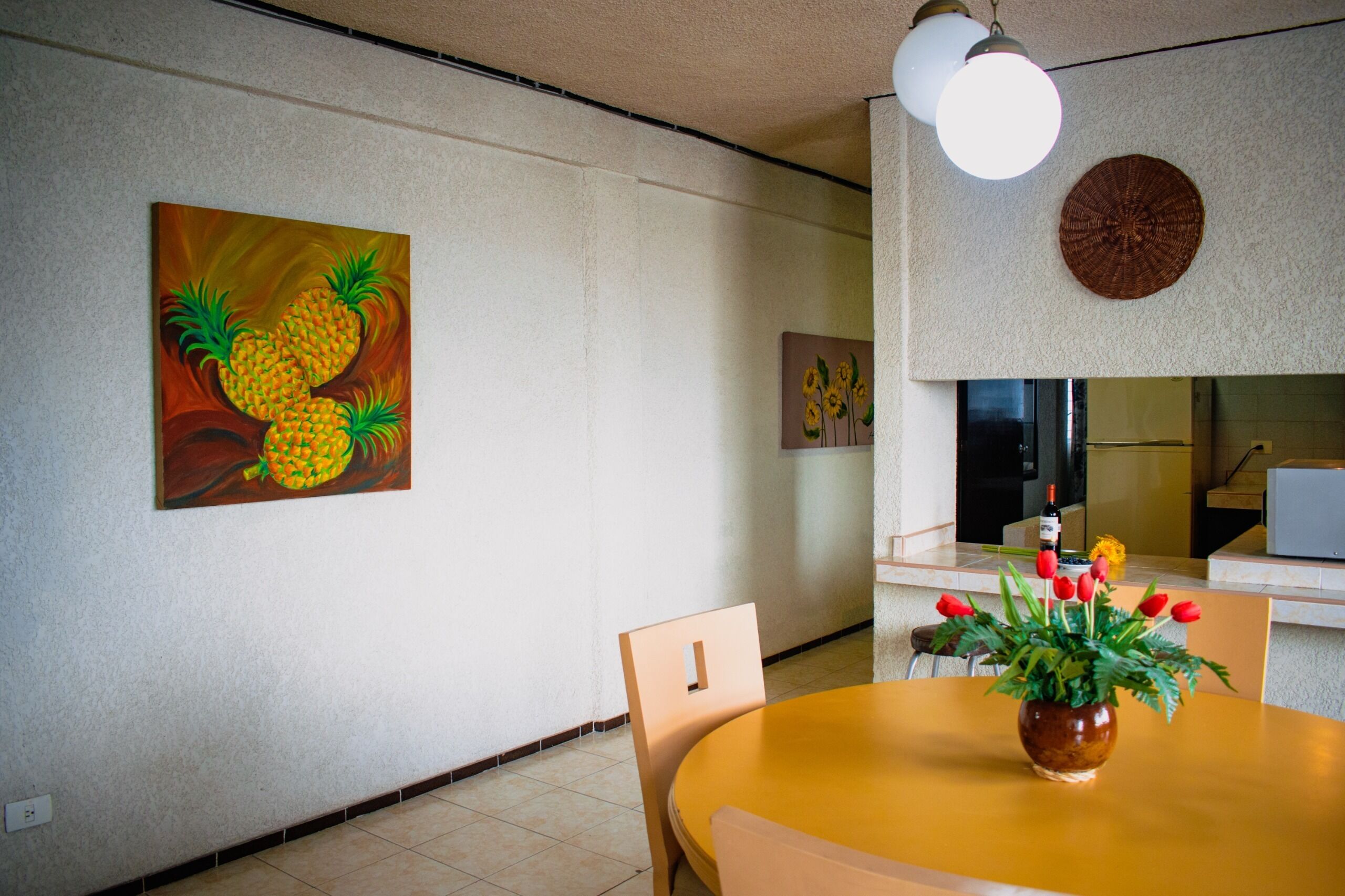 A dining area with a round table, chairs, a vase with red flowers, and wall art depicting pineapples and sunflowers.