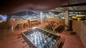 A rooftop bar with a foosball table, seating area, and a view of a cityscape at night.