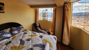 A bedroom with a bed, a chair, a window with curtains, and a view of a cityscape.