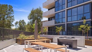 Une terrasse extérieure dotée de bancs en bois et d’un espace barbecue, le tout entouré d’un bâtiment moderne aux grandes fenêtres.