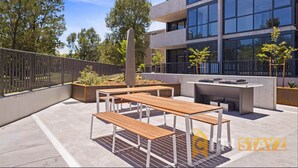 Une terrasse extérieure avec des bancs en bois et un espace barbecue, entourée d’un bâtiment moderne doté de grandes fenêtres.