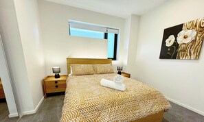 A neatly made bed with patterned bedding, two bedside lamps, a small wooden nightstand, a wall-mounted air conditioning unit, and a floral artwork on the wall. - Lavish in Lyneham - 2BD 1BTH Apt (Lyneham)