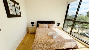 Une chambre avec une grande fenêtre, un lit recouvert d’une literie à motifs, deux tables de chevet avec des lampes et un tableau accroché au mur.