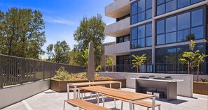 Une terrasse extérieure avec des bancs en bois et un espace barbecue, entourée d’un bâtiment moderne doté de grandes fenêtres.