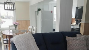 Una sala moderna con un sofá azul oscuro, una manta blanca y almohadones estampados. Se ve un área de comedor con una mesa y sillas de madera al fondo.