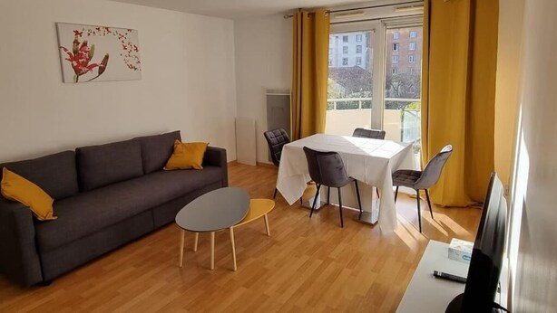 Ein Wohnzimmer mit einer Couch, einem Esstisch und Stühlen. Ein Fenster mit Vorhängen und ein Bild an der Wand sind ebenfalls zu sehen.