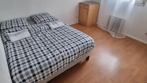 Ein Schlafzimmer mit einem Bett, das mit einer karierten Decke bezogen ist, einer kleinen Holzkommode und einem Fenster mit durchsichtigen Vorhängen.