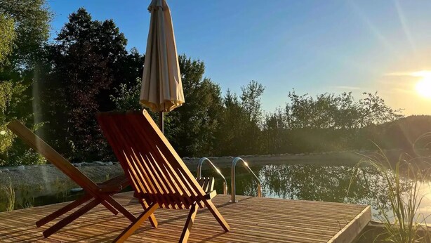 En træterrasse med en rød liggestol og en beige parasol, med udsigt over et roligt vandareal og træer.
