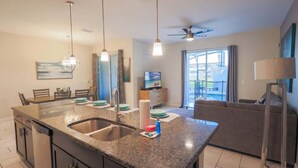 A modern kitchen with granite countertops, a double sink, and pendant lighting. There is a dining area with a table and chairs, and a living room with a sofa and a TV. - Magical Townhome private pool in Storey Lake (Kissimmee)
