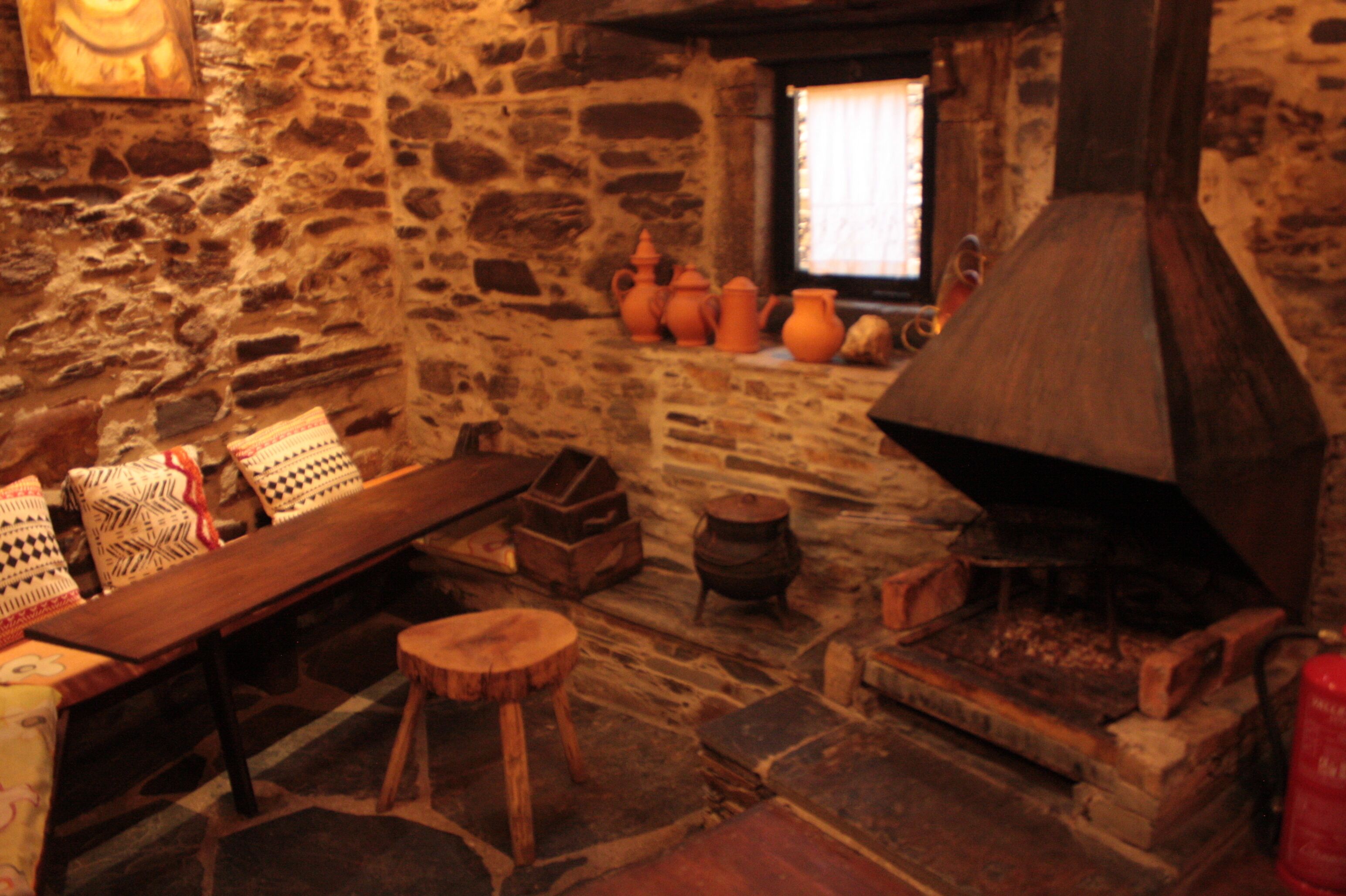 Una habitación rústica con paredes de piedra, una mesa de madera, bancos y un horno de leña tradicional.