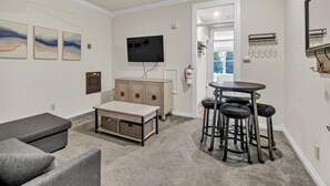 Una sala de estar con un sofá, un reposapiés y una pequeña mesa con taburetes. Hay un televisor montado en la pared, un ventilador de techo y una puerta que da a otra habitación.