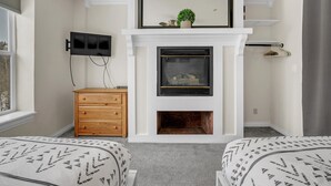 Un dormitorio con chimenea, televisor montado en la pared, cómoda de madera y dos camas con ropa de cama estampada.