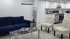 Moderni olohuone, jossa on sininen sohva, sohvapöytä ja kuviolliset tuolit. Taustalla näkyy keittiö, jossa on valkoiset kaapit ja nykyaikaiset kodinkoneet.