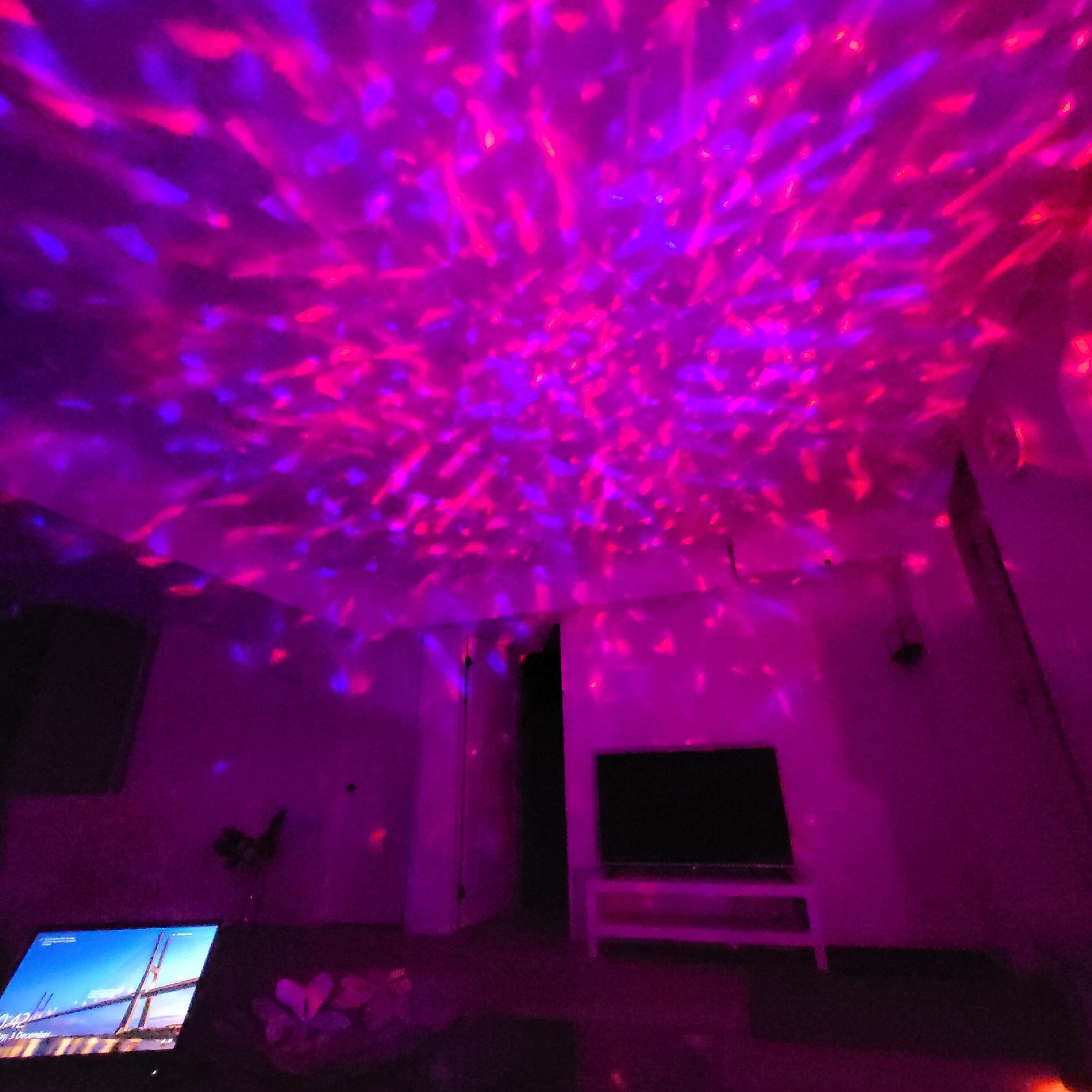 Una habitación con proyección en el techo de luces rosadas y azules, un televisor y una banca.