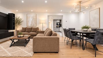 Een moderne woonkamer met een zithoek, een eettafel met stoelen, een aan de muur bevestigde televisie en een keuken op de achtergrond.