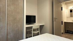 Une chambre moderne avec un lit, un bureau doté d’un téléviseur, une chaise et une salle de bain visible à travers une porte ouverte.
