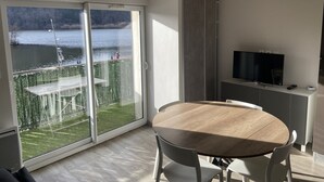 Une chambre moderne dotée d’une grande porte-fenêtre coulissante donnant sur un balcon avec vue sur un lac et des arbres. On y trouve une table ronde en bois avec des chaises, un téléviseur posé sur un meuble et un petit placard.
