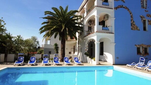 A pool with blue chairs, a palm tree, and a building with white and blue walls. - [Cedro] Apartment with swimming pool (Orosei)