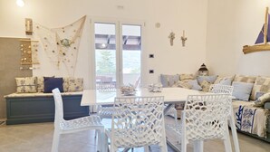 A dining area with a white table and chairs, a bench with cushions, and a view through a glass door. - [Cedro] Apartment with swimming pool (Orosei)