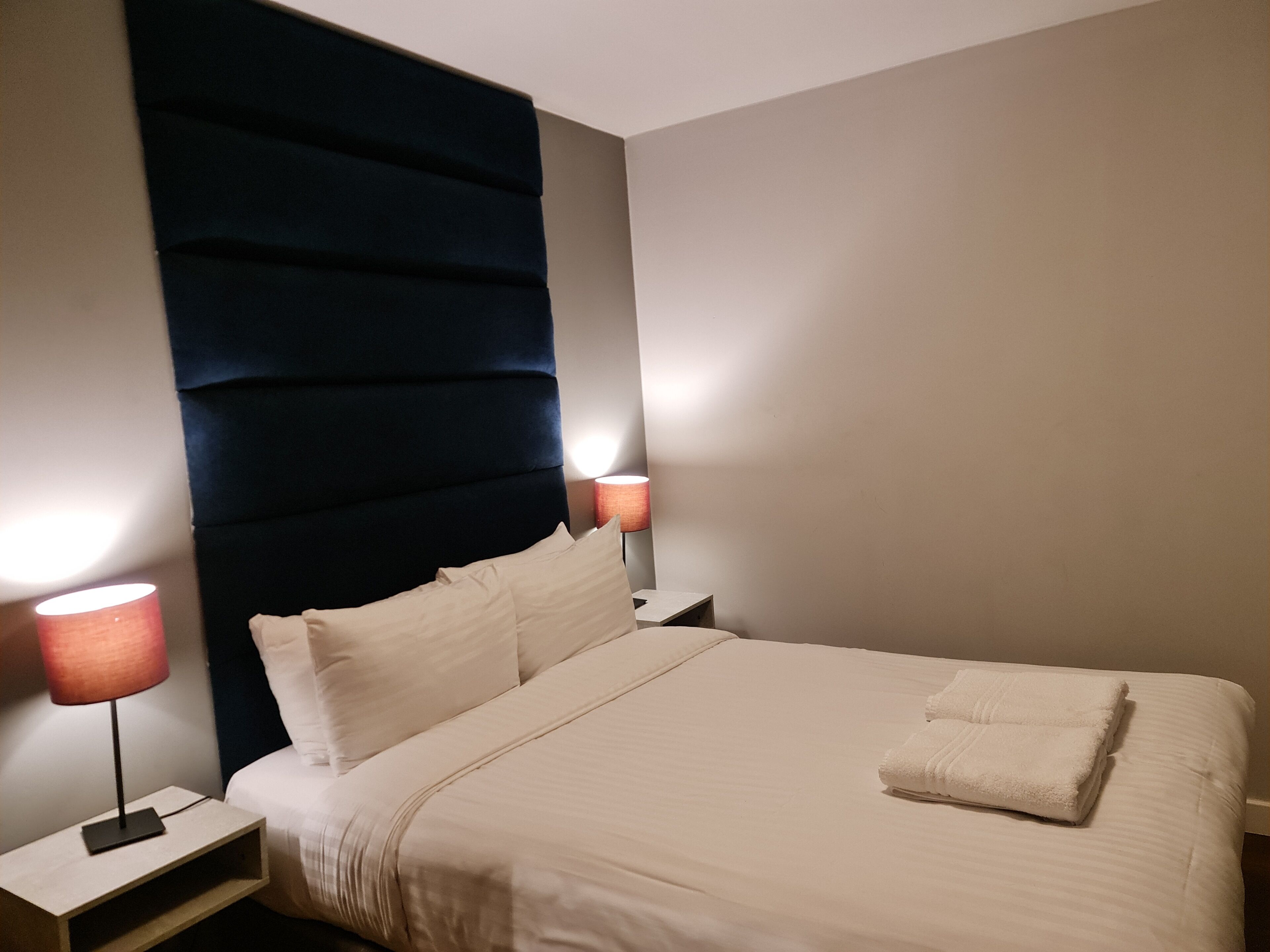A neatly made bed with white linens and a folded towel, a dark upholstered headboard, two bedside tables with lamps, and a plain wall.