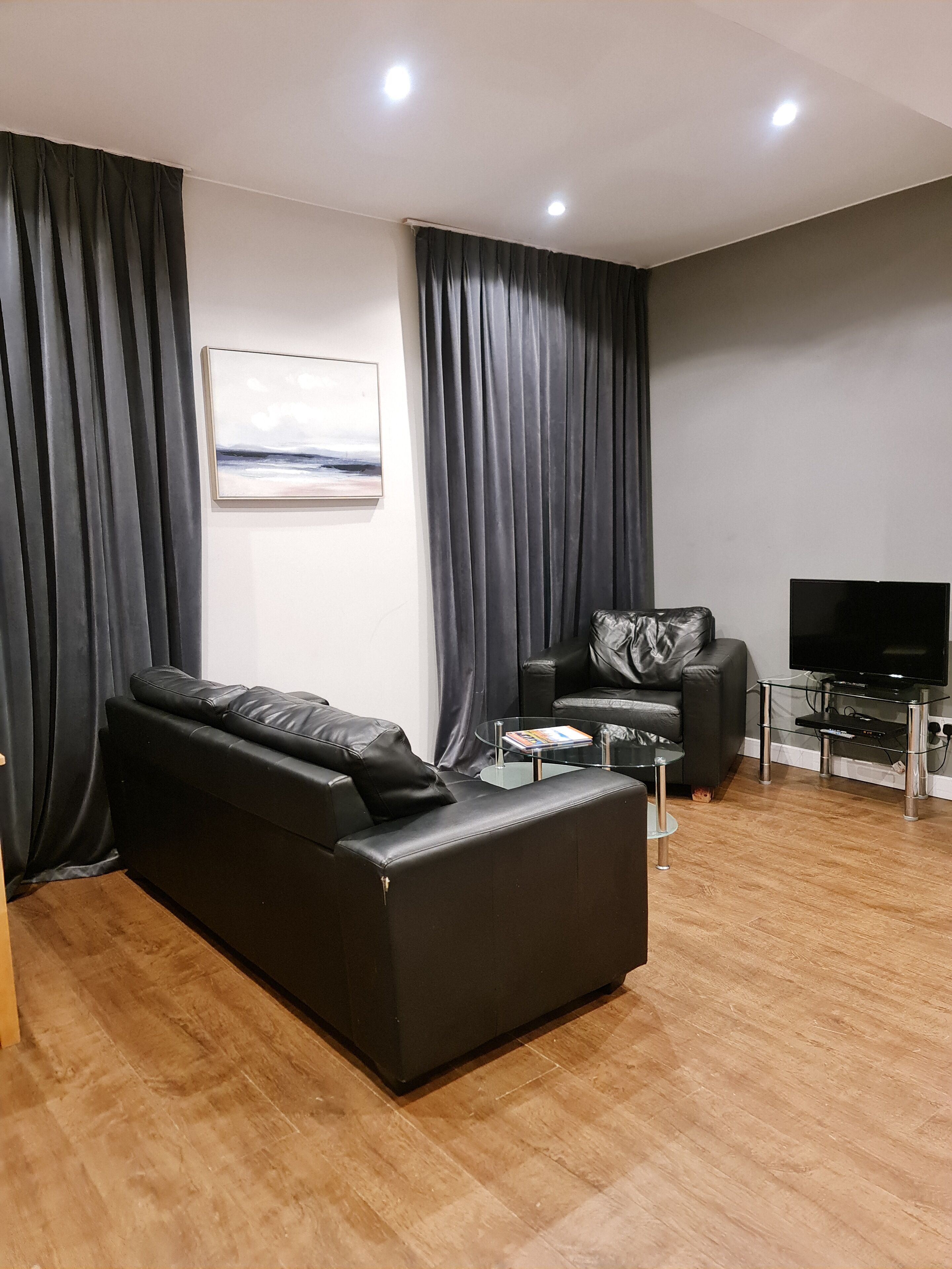 Een moderne woonkamer met een zwarte leren bank, een glazen salontafel, een flatscreen televisie en donkere gordijnen.